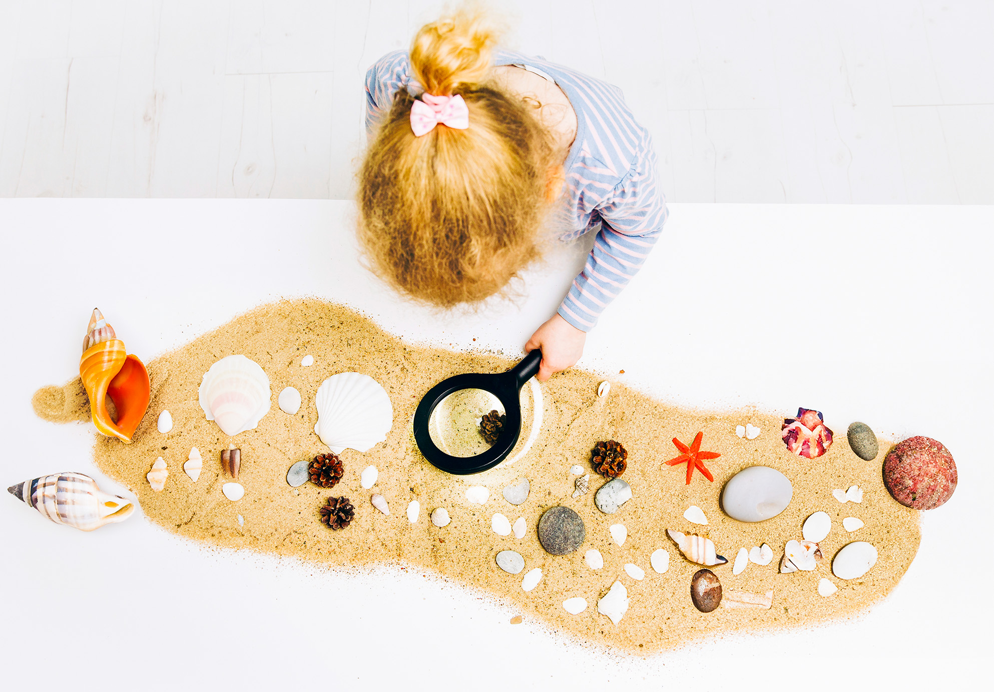 Overhead view of a young child, using a magnifying glass to inspect sand and seashells